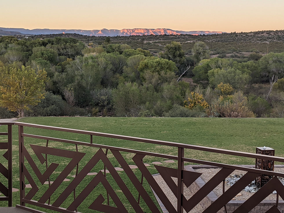 Distant cliffs in sunset colors, as seen from the River Room balcony. A field of green grass is below, with Cottonwood trees dominating the mid-range view.