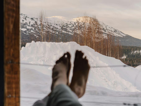 A cozy winter scene viewed from indoors, with someone resting their legs while looking out at snowy mountains and bare trees — symbolizing calm, rest, and gentle winter self-care.