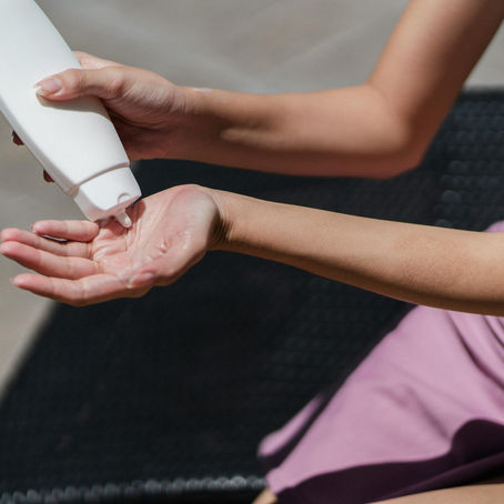 A woman wearing a pink skirt and white bikini top applies sunscreen from a bottle into her left palm. The image focuses on her arms and hands, capturing the ritual of sun protection under bright sunshine.