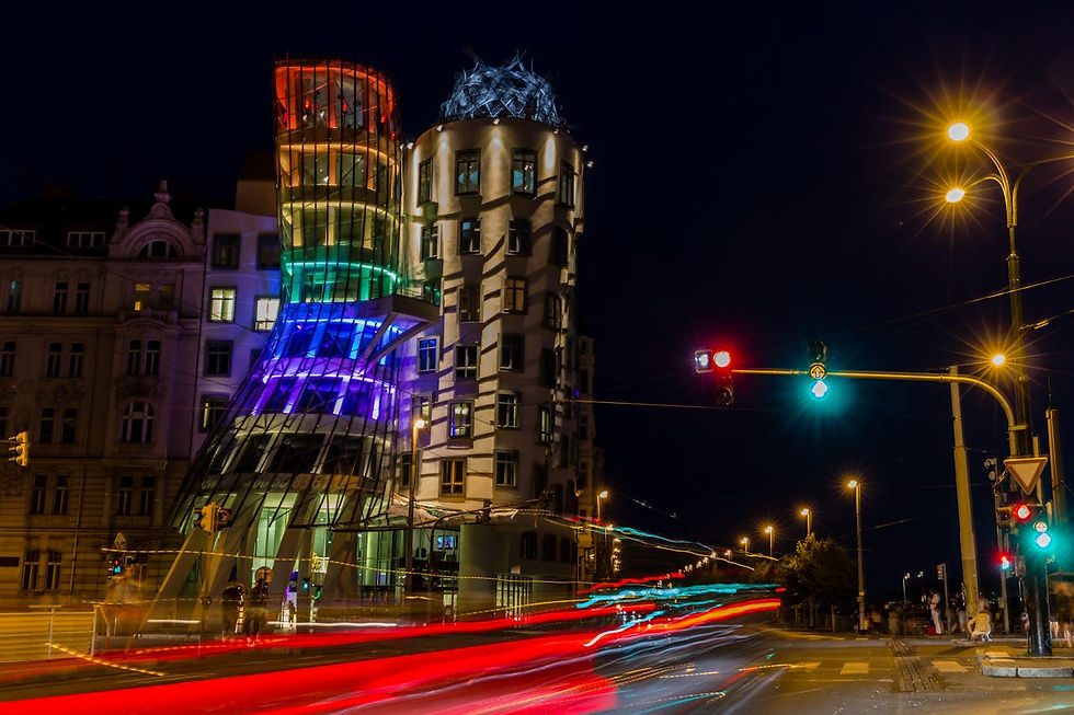 Nighttime photo of Prague’s Dancing House (Tančící dům) illuminated with colorful lights, with long-exposure car light trails on a city street and warm street lamps, capturing an energetic Prague city-night atmosphere for a stag weekend comparison article.