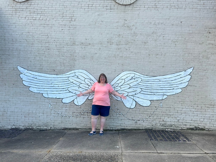 Woman with arms outstretched to each side while standing in front of a painting of large white wings on the side of a brick building