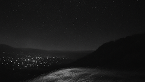 A hillside of windswept grass glows faintly in the night, with a vast starry sky and twinkling city lights in the distance.