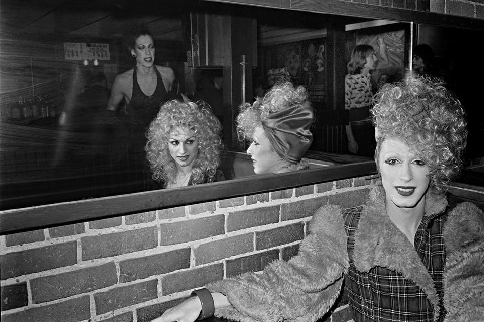 Stylish female impersonator seated in booth with friends reflected in mirror.