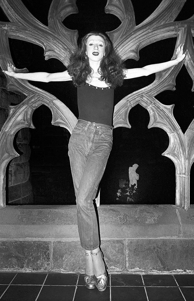 Drag queen with jeans and tight shirt, posing at church courtyard site.