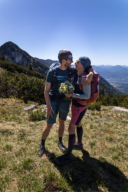 Ein Brautpaar in Bergkleidung mit Rucksäcken und Brautstrauß umarmt sich auf einem sonnigen Berggipfel nach einer Wanderhochzeit vor beeindruckender Alpenkulisse. 