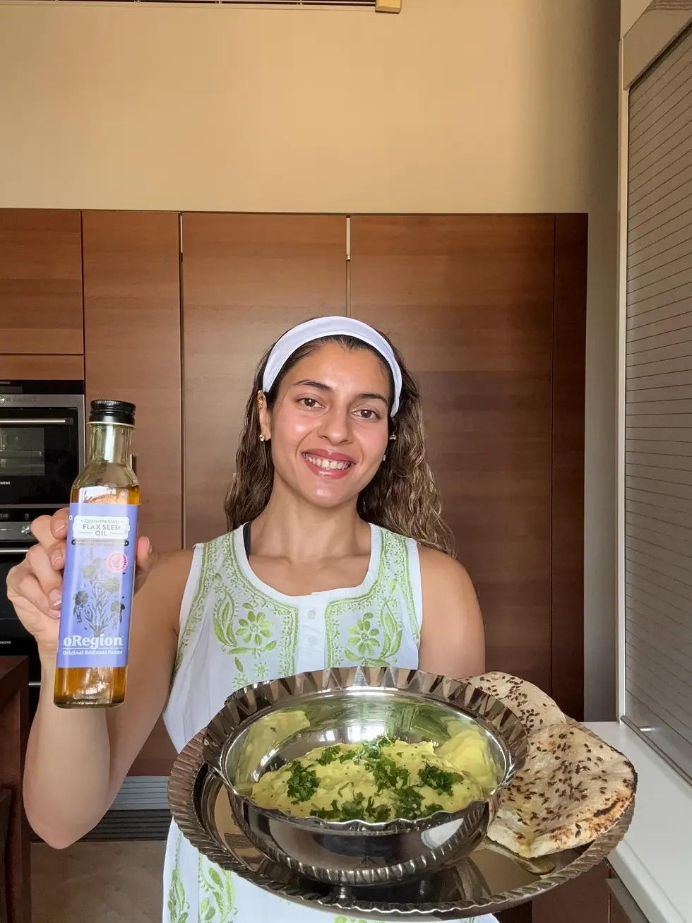 Smiling woman in a kitchen holds flaxseed oil and a tray of food with flatbread and a green garnished dish.