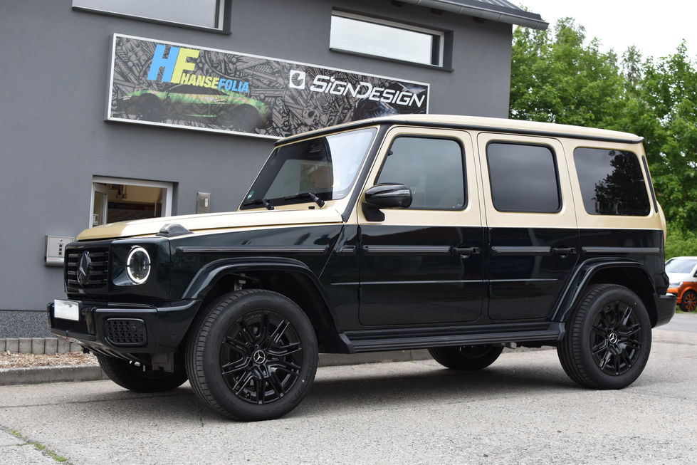 Schrägansicht einer Mercedes G-Klasse in einer Zwei-Farben-Folierung: Oben sandfarben, unten schwarzglänzend. Das Fahrzeug steht vor einem Firmengebäude von "SignDesign" und "Hansefolia". Die Felgen sind komplett schwarz.