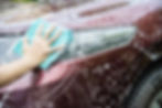 Hand wiping soapy foam off a maroon car’s headlight with a blue cloth during a car wash.