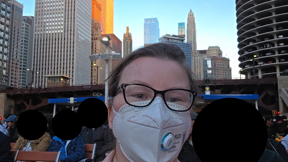 Masked person with glasses takes a selfie on an outdoor boat deck, with downtown skyscrapers and seated passengers behind them.