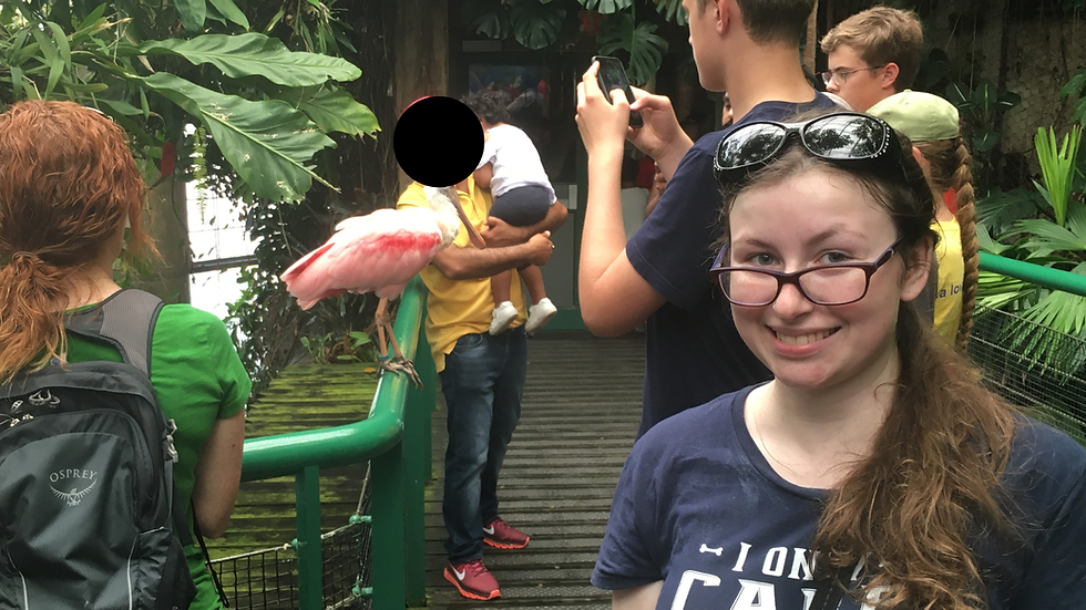Smiling girl in glasses at a tropical bird exhibit, with a pink spoonbill on a railing and visitors taking photos