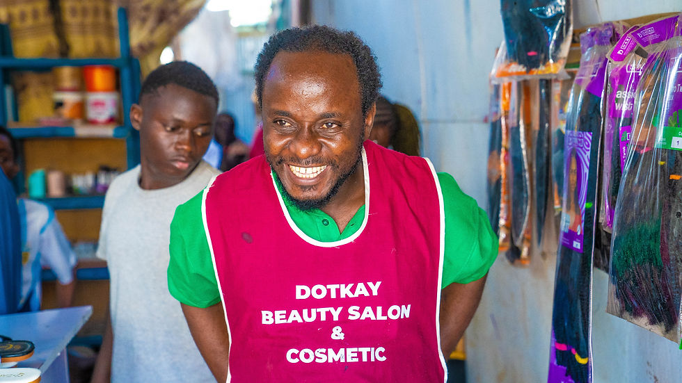 Smiling man in a magenta apron labeled DOTKAY BEAUTY SALON & COSMETIC stands in a small shop, with another man behind him.