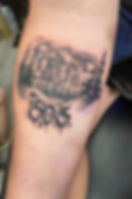 Tattoo of a city skyline with mountains and trees on skin. "303" in bold graffiti-style below. Monochrome design.