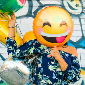 Eine Frau in geblümter Bluse, die ihr Gesicht hinter einem lachenden Emoji-Ballon versteckt