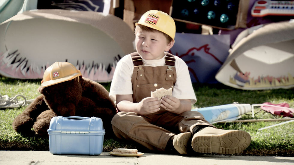 Young boy in overalls eating a sandwich, sitting next to a teddy bear and blue lunch pale
