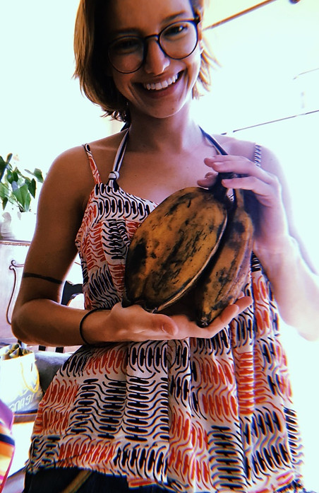 Mulher branca, cabelos castanhos, sorrindo, usando óculos preto, blusa de alça estilo bata, branca com detalhes em preto e laranja, segurando um cacho de bananas à frente do seu tronco, ao fundo, vaso branco de cerâmica com planta verde sobre um móvel antigo