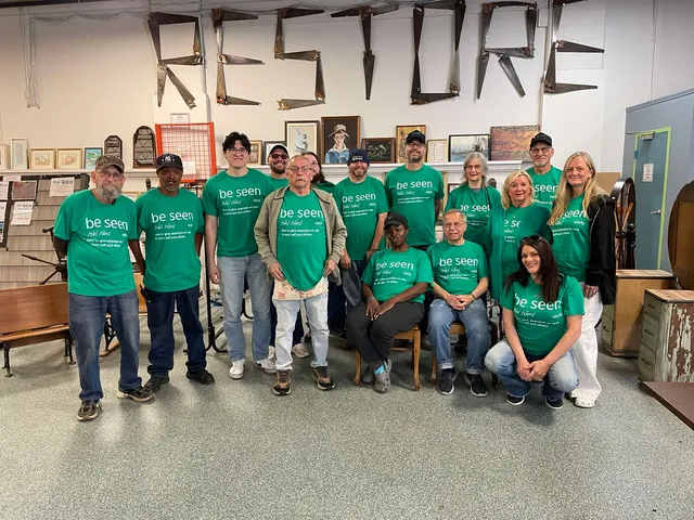 Group photo of Lima ReStore staff and volunteers wearing green “be seen” Mental Health Awareness shirts inside the store beneath a large “RESTORE” sign made from reclaimed materials.