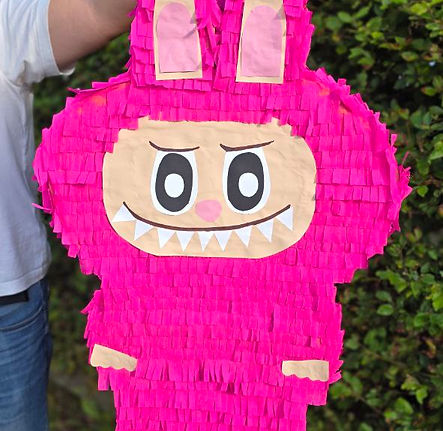 En rosa monster-piñata holdes opp foran en grønn hekk.