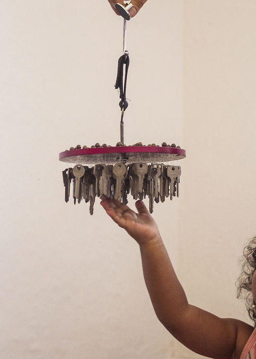 Detalhe da mãe e braço de uma criança pequena negra de pele clara, tocando, com a ponta dos dedos, um chavilhão pendurado acima de sua mão. Vivência musical, musicalização, musicoterapia, intervenção precoce, neurorreabilitação, terapia com música, intervenção a tempo - T21 trissomia Sindrome de down, TEA transtorno do espectro autista, paralisia cerebral