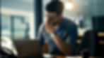 A man in glasses, wearing a blue shirt, appears stressed while working on a laptop in an office with dim lighting. A cup and notes are nearby.