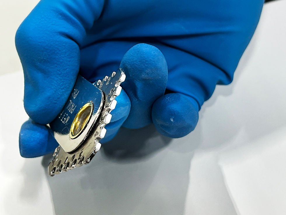 Blue-gloved hand holding a shiny, metal wet film gauge with white paint on the teeth. The background is white, highlighting the tool's details.