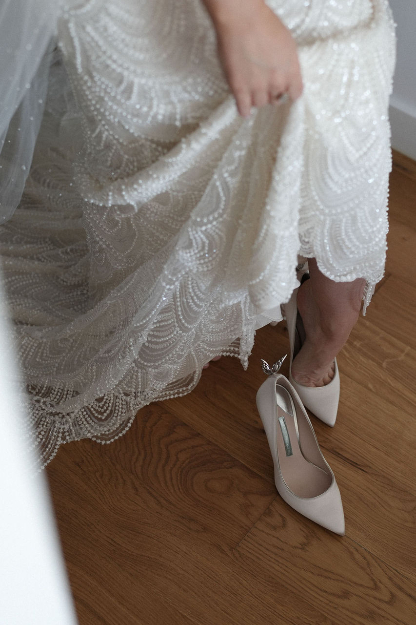 A close-up of April's intricately beaded wedding dress, next to her bridal shoes on a wooden floor.
