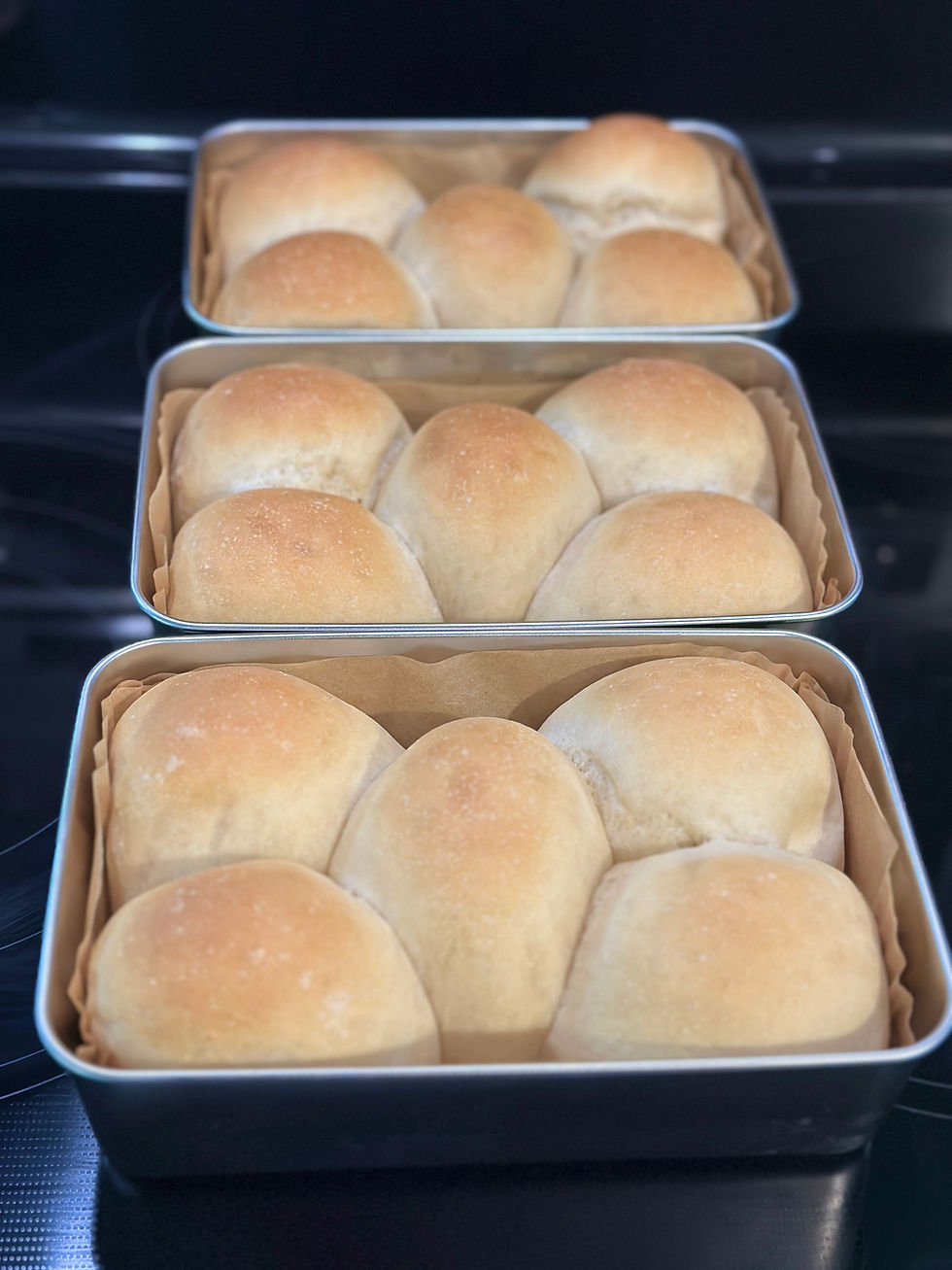 Three pans of golden brown sourdough Hawaiian rolls fresh from the oven sitting on a glass stovetop