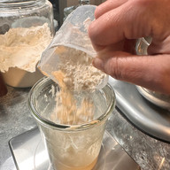 Flour being added by weight to a sourdough starter in a Weck jar on a kitchen scale during the feeding process