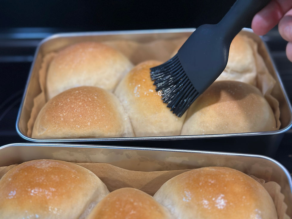 Black silicone pastry brush applying melted butter to the top of a golden sourdough Hawaiian roll directly in the loaf pan