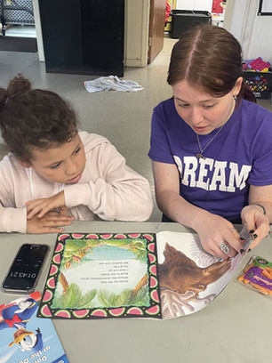 A person wearing a DREAM shirt points at words in a picture book reading to a child. A phone timer records and is placed on the table next to the book.