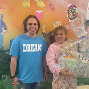 Two smiling people stand in front of a colorful mural. On the left, a man wears a blue "DREAM" shirt; the girl on the left holds a gift basket wrapped in cellophane.