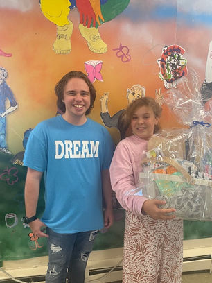 Two smiling people stand in front of a colorful mural. On the left, a man wears a blue "DREAM" shirt; the girl on the left holds a gift basket wrapped in cellophane.