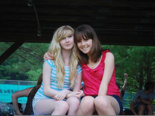 Two smiling people sitting together under a wooden shelter, one in a striped shirt, the other in pink. Background shows trees and people.