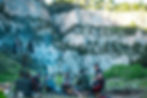 Group of people sitting around a campfire in front of rocky cliffs and green trees, with flags visible. The mood is relaxed and sociable.