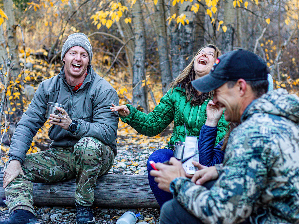Three people in warm clothing laugh joyfully while holding mugs in a forest with yellow leaves, evoking a cheerful, autumnal mood.