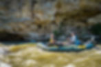 Two people in a raft navigate turbulent waters beside a rocky cliff. The bright blue raft contrasts with the earthy stone background.