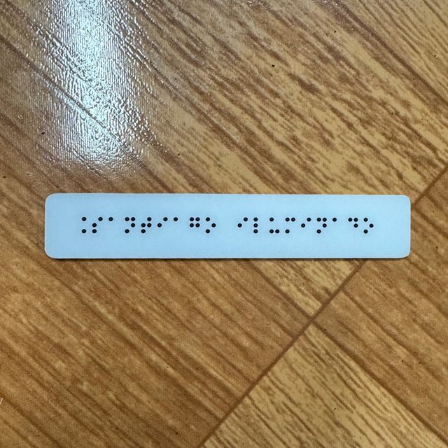 Placa alargada y angosta de acrílico blanco con texto solo en braille negro en relieve