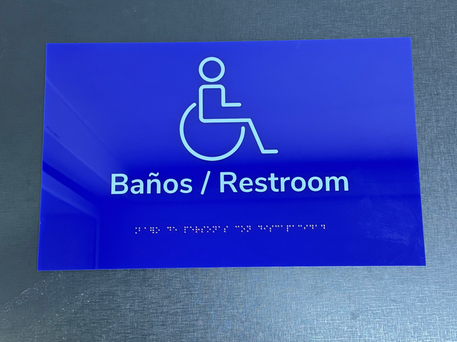 Placa de acrílico azul "Baños / Restroom" con símbolo de silla de ruedas en celeste y braille en relieve