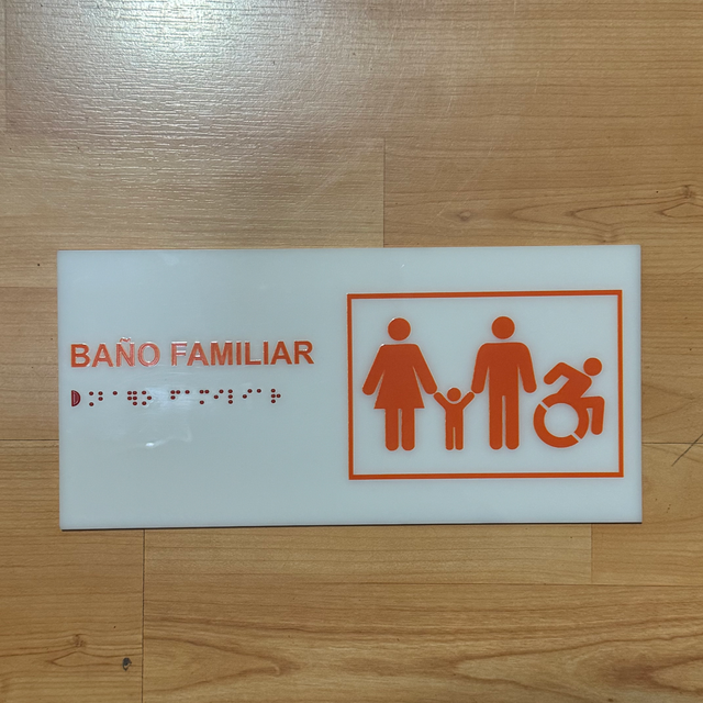 Placa de acrílico blanco "Baño familiar" en naranja, con pictograma de familia y silla de ruedas en recuadro, y braille