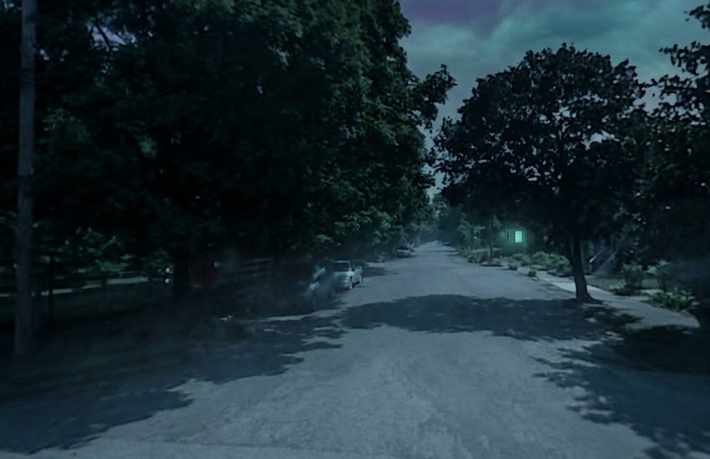 A dark, tree-lined residential street at night, enveloped in shadows with dark purple storm clouds overhead and a solitary, eerie green light glowing from a house in the distance.