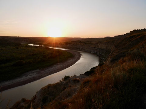 Golden sunset glowing over the curving Little Missouri River framed by dark badlands cliffs.