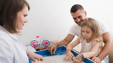 young girl accompanied by her father plays in a sand play therapy observed by a female therapist