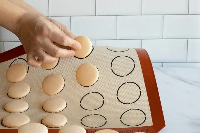 Hands lifting beige macarons from a reusable silicone baking mats with circle guides on a marble counter. Background features white subway tiles.