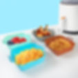 The image shows four silicone air fryer liners in different colors (blue, green, orange, and grey), each containing different food items. The blue liner holds a mix of blueberries and raspberries, the green contains chicken drumsticks, the orange is filled with tater tots, and the grey one has french fries. In the background, there is a white air fryer appliance. These liners are reusable alternatives to disposable parchment paper or foil, suggesting an eco-friendly option for cooking.