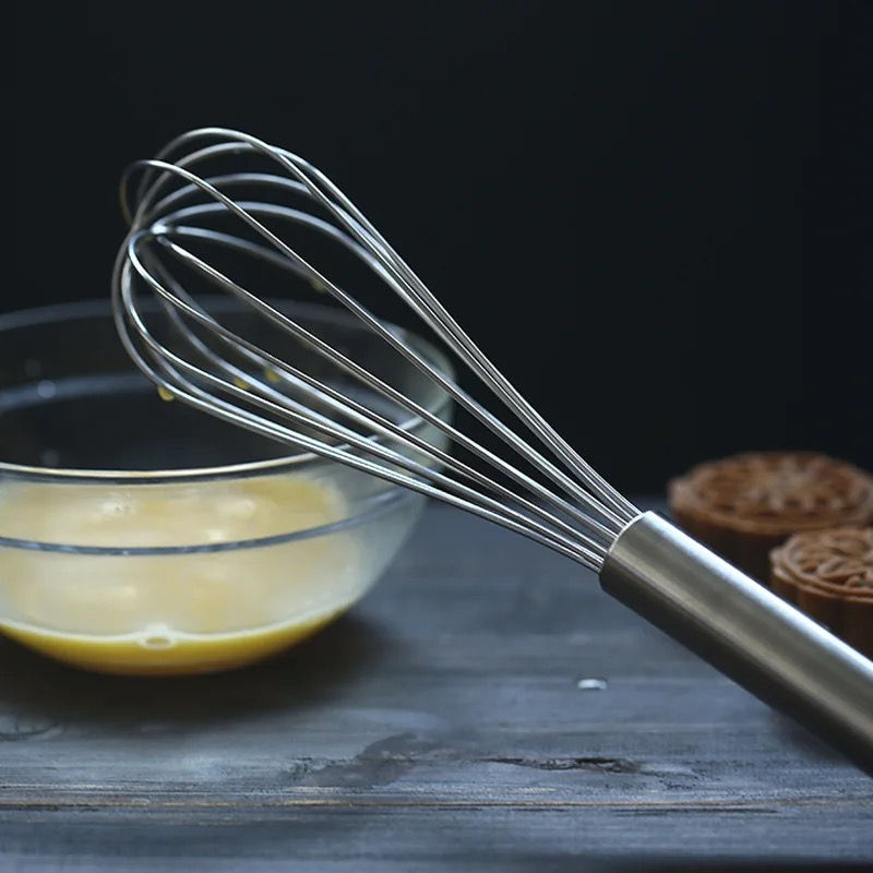 Thumbnail: A stainless steel whisk from Bakecraft Tools with a slender handle and a series of looping wires forming the head, positioned in the foreground on a dark surface. In the background, there is a glass bowl containing a yellow mixture, likely batter, and to the right are partially visible round baked goods with intricate designs on them. The focus on the whisk against the dark backdrop highlights its sleek design and suggests its use in baking preparation.