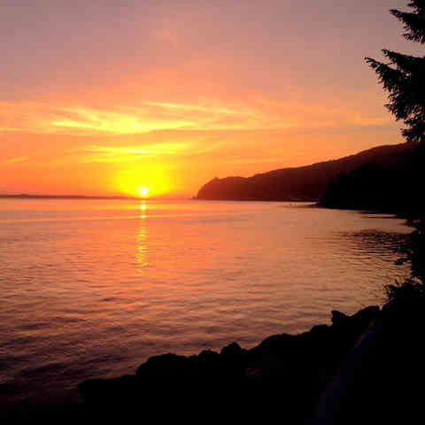 The sun is setting over the horrison of the edge of a cresent shaped cove and the sun is reflected in the water and light up the orange, pink and purple sky with golden light.