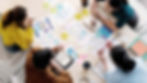Four coworkers brainstorm UX designs around a table with phone mockups, sticky notes, notebooks, and colorful charts.