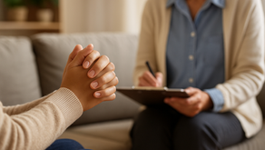 "A close-up of a person’s clasped hands during a counseling session in Sarasota, symbolizing faith and emotional reflection, with a compassionate therapist blurred in the background taking notes in a calm, softly lit office."