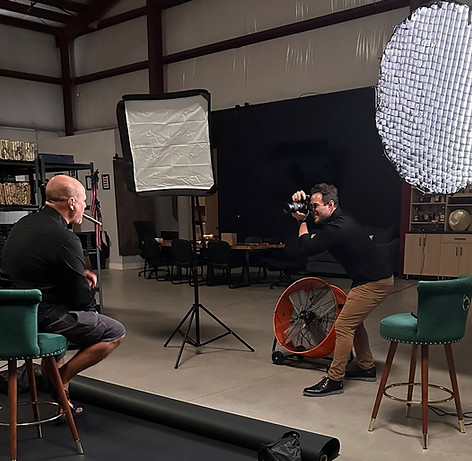 Ledford Productions - Photography setup in a large room with a high ceiling, featuring a seated man under a softbox light, a photographer behind him, and scattered equipment on a concrete floor.