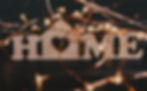 Wooden letters spelling "HOME" with a heart-shaped cutout in the "O" are surrounded by warm, glowing string lights against a dark background. Cozy ambiance.