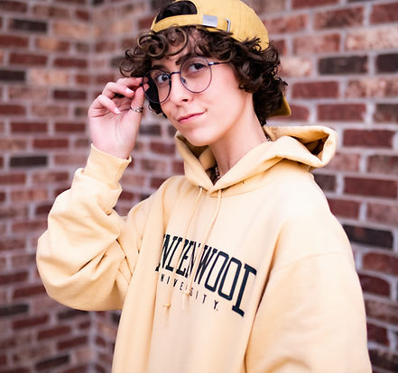 Sammi Reise- Person with short curly dark hair and glasses wearing a yellow hoodie and backwards baseball cap.